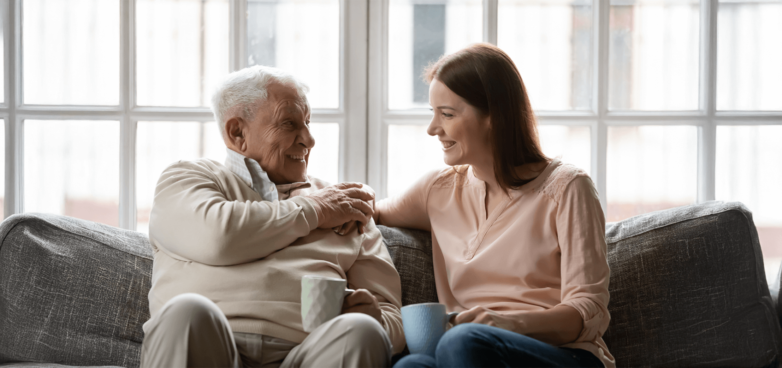 Elderly man and young woman smiling while sitting on a sofa, holding mugs