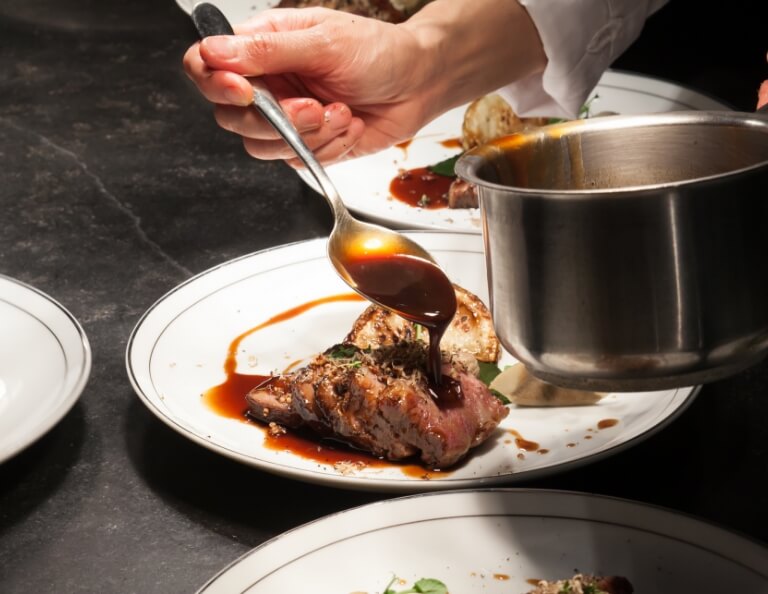 Hand pouring sauce over a steak dish in a professional kitchen setting.