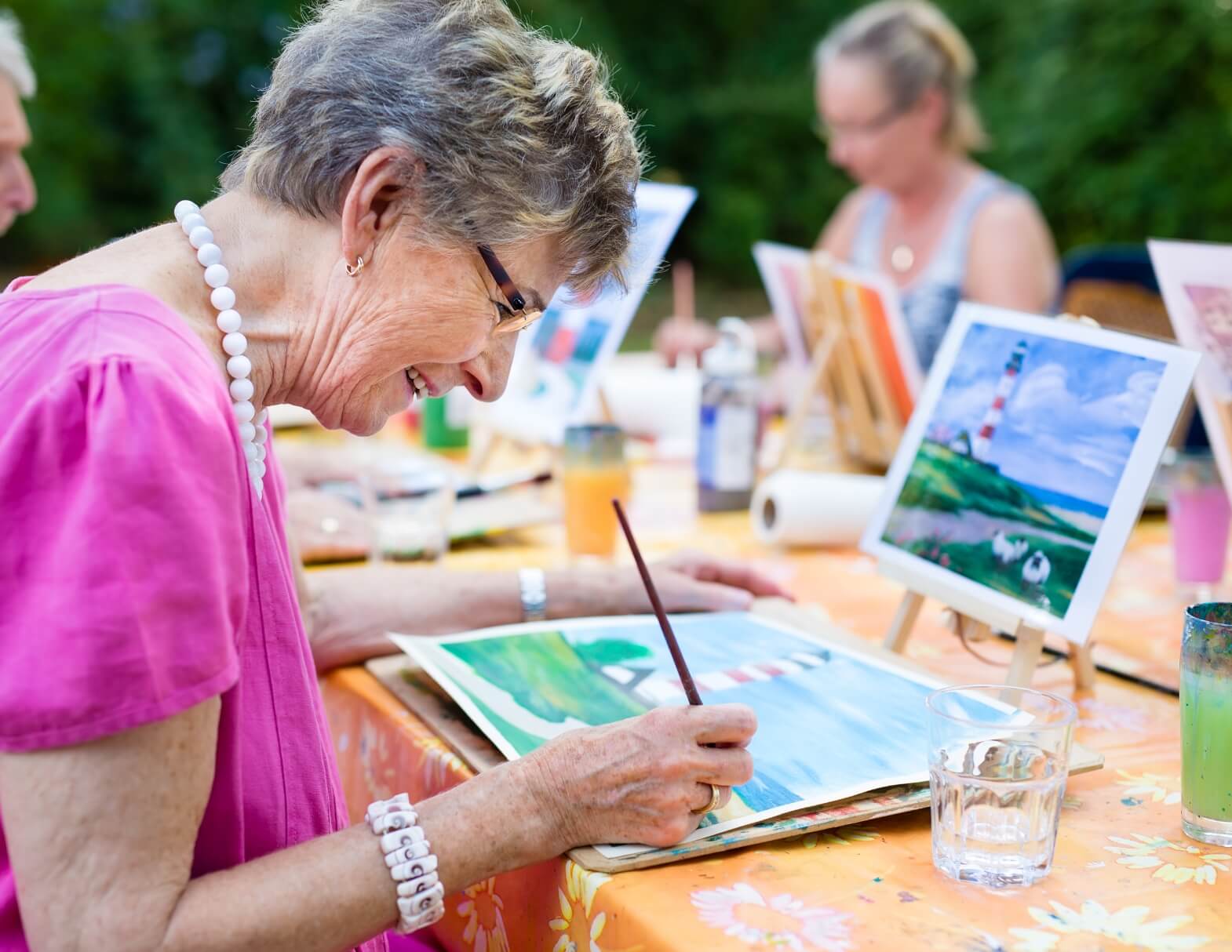 Senior woman in pink dress enjoys painting outdoors with friends at an art class.