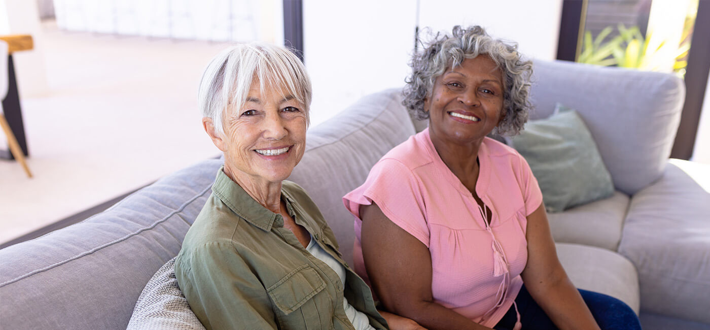 Two senior women sit on couch smiling while looking up