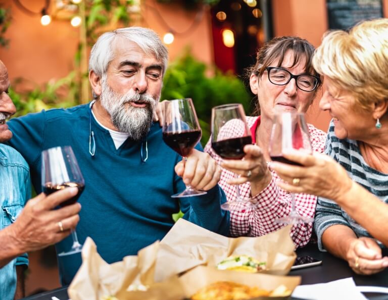 Group of seniors toasting with red wine glasses outdoors at a dining table.