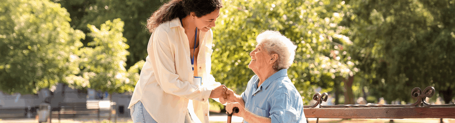 Senior woman sits on an outdoor bench smiling up at younger female caretaker
