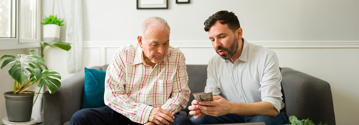 Senior man and younger man sit on a couch together and review finances